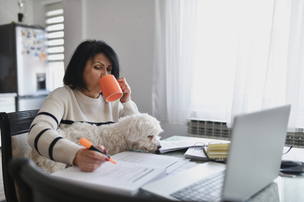 Teacher Drinking Coffee While Grading Exams at Home with Curious Dog Sitting on Her Lap. Self-care. Wellness. Well-being.