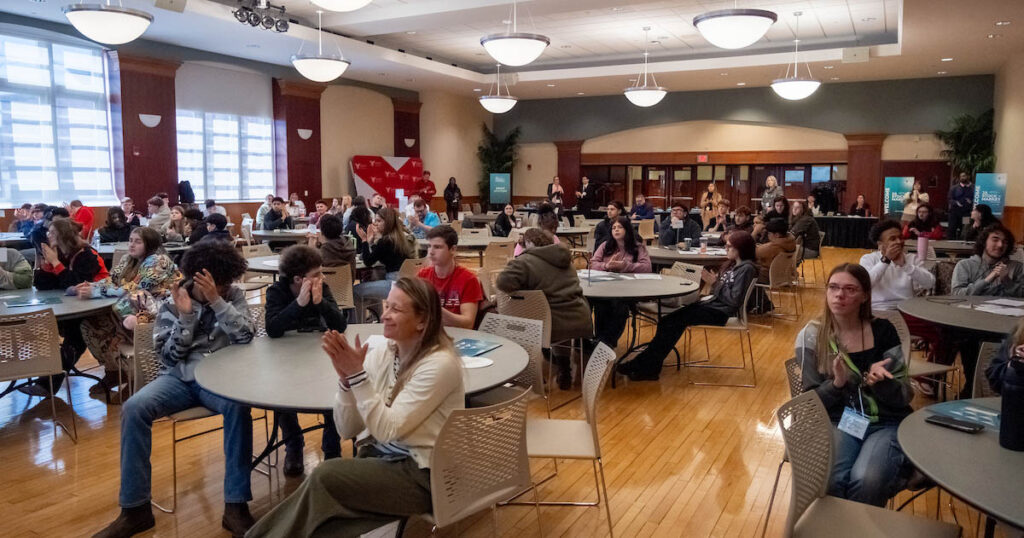Bells, Trades, and Big Decisions: High School Students Test Their Investing Skills at Ball State People seated at various tables in a large, well-lit room smile and clap at an event.