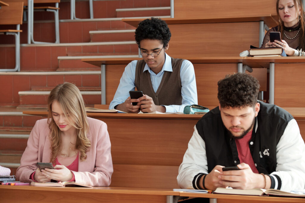 Group of contemporary young intercultural students in casualwear sitting by desks in lecture hall and scrolling in smartphones at break