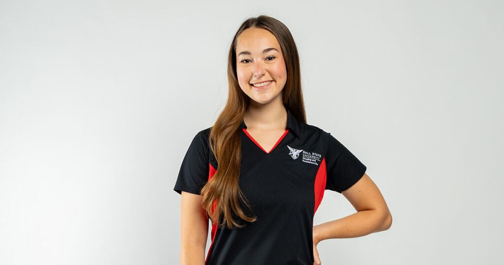 A young woman with long brown hair wearing a black and red Ball State collared shirt, posing and smiling in front of a white backdrop.