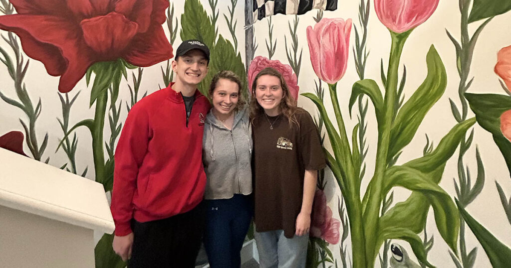 Three students stand and smile in front of an indoor mural that displays various large pink and red flowers with greenery