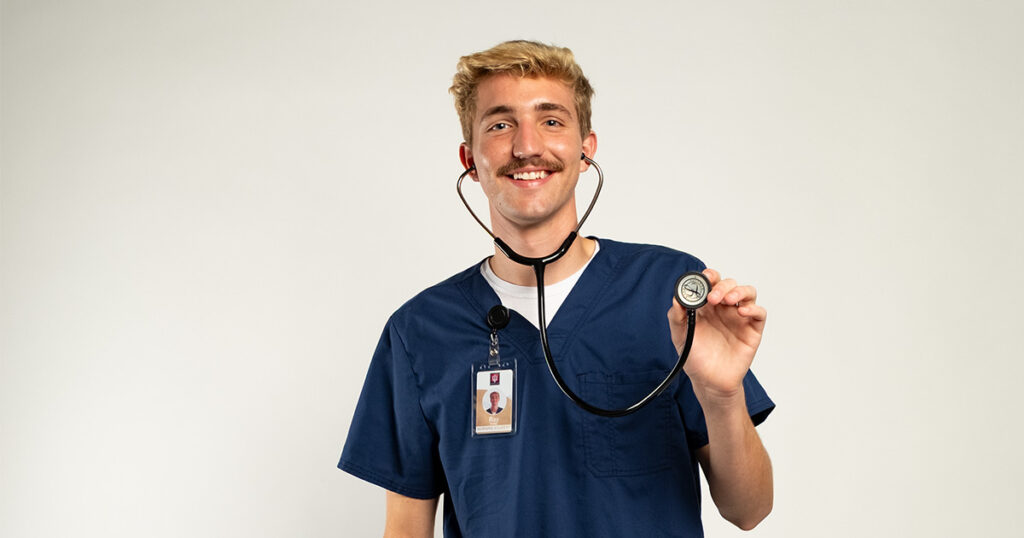 From cautious to caring—advancing in compassionate nursing practice. A young man with short blonde hair wearing a blue scrub top and a stethoscope, posing and smiling in front of a white backdrop.