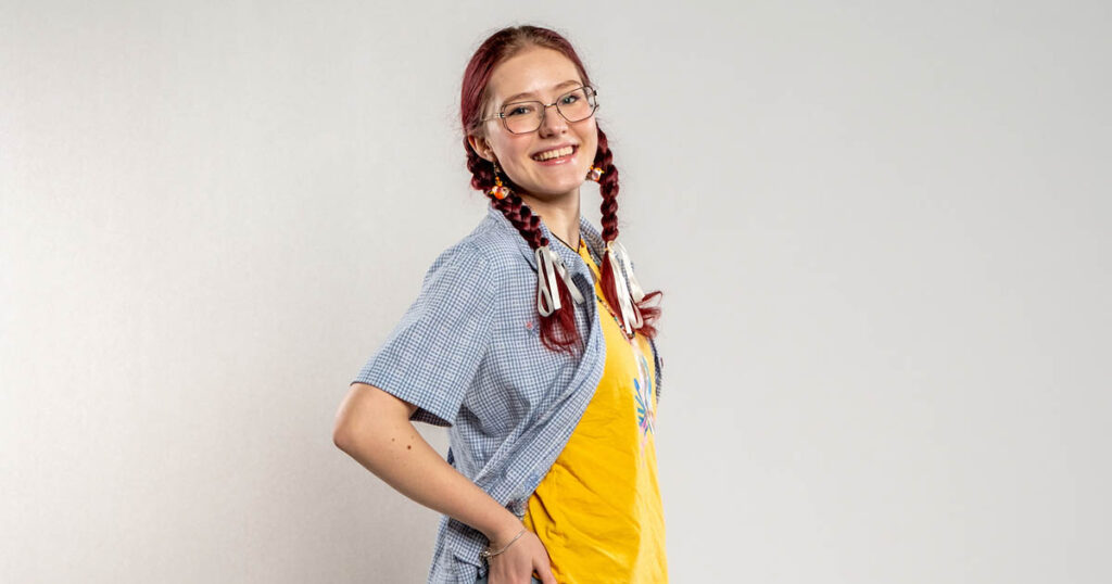 Portrait of Viktoriia Burdiad, wearing glasses, a blue top, and yellow undershirt while smiling at the camera with a hand in her pocket.