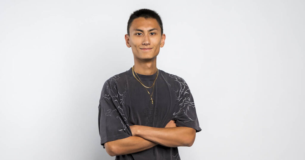 Portrait of Reo Horie, wearing a black t-shirt and gold necklace while smiling and crossing his arms.