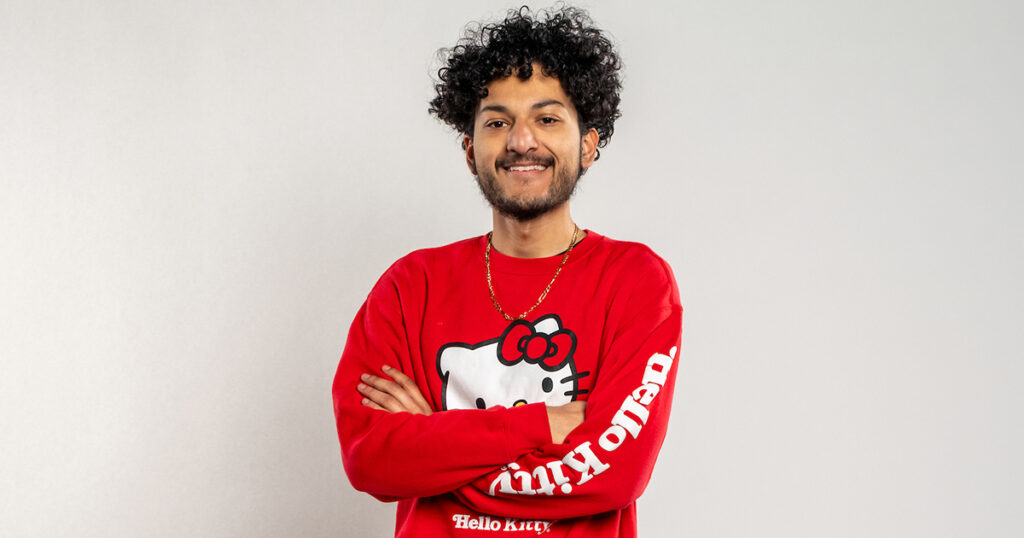 Portrait of Angel Esquivel Vazquez, with dark, curly hair smiling while crossing his arms and wearing a red long-sleeve shirt.