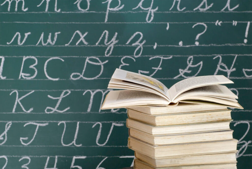 Image of an open book atop a stack of books in front of a blackboard with the alphabet written on it in cursive in chalk.