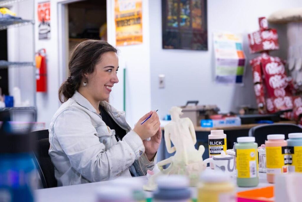 Art & Design Goes Global, Sparks Growth Student in Shenandoah University's Art & Design program smiles and looks away from the camera as works on a small sculpture of a seated human figure.