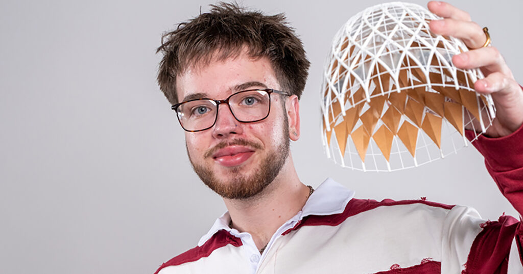 Brodie Budzinski, a young man with short brown hair, brown facial hair, wearing glasses and a red and white striped sweatshirt and holding up a weaved 3D model