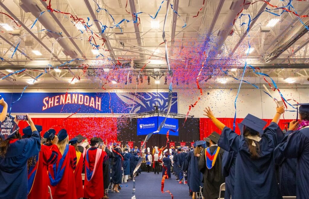 Multicolored streamers fall from the ceiling of the Wilkins Athletics & Events Center at a University Commencement. Graduates in blue caps and gowns and red doctoral robes celebrate.