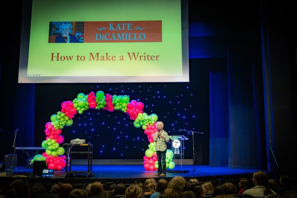 Author Kate DiCamillo in front of a pink and green balloon arch and a slide that says "How to Make a Writer" on stage at Shenandoah University's Ohrstrom-Bryant Theatre in 2026 for the university's Children's Literature Conference's Rally for Reading.